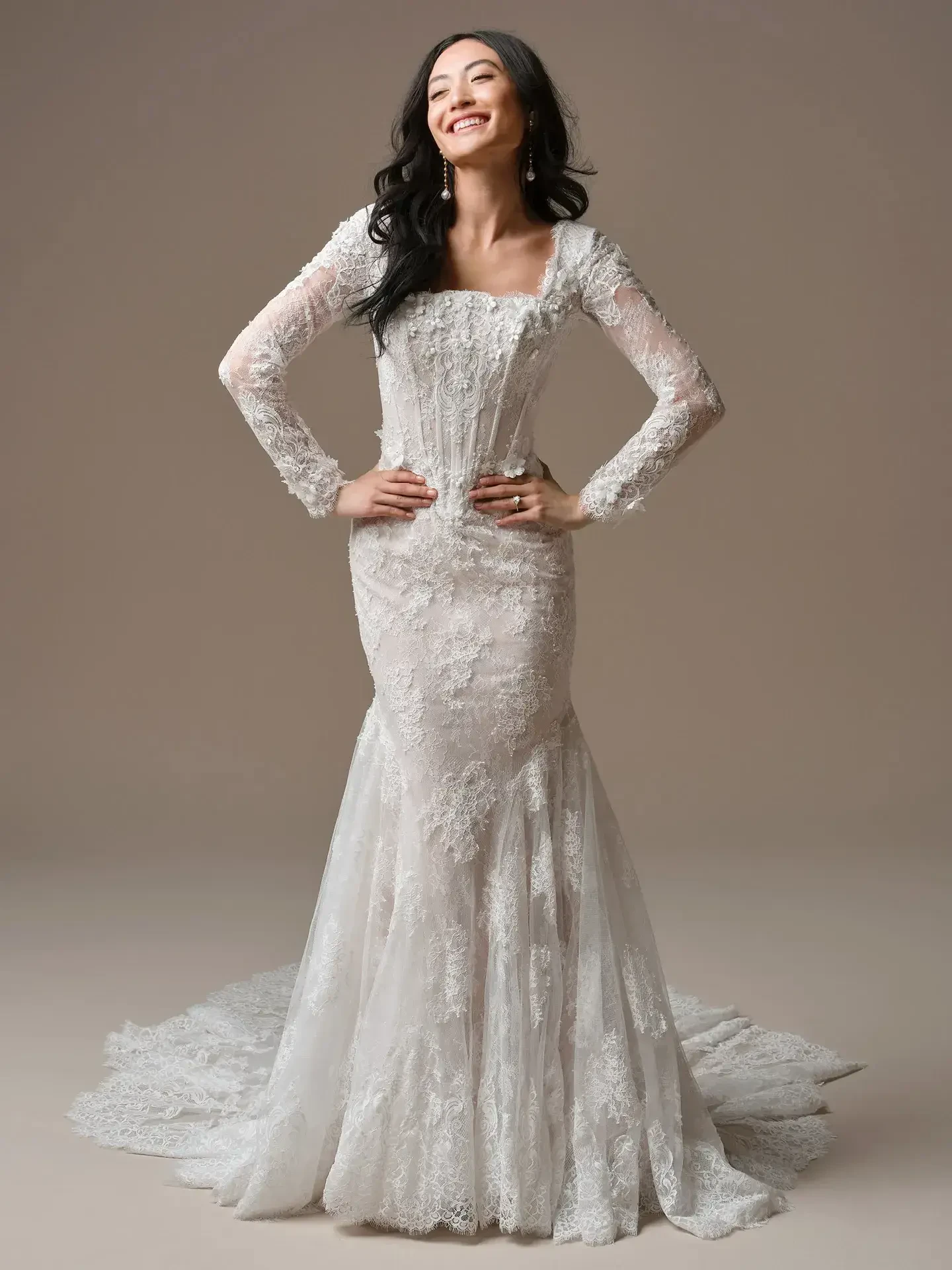 Woman in a lace mermaid wedding dress stands confidently with hands on hips, smiling. The dress features long sleeves and an elegant train.