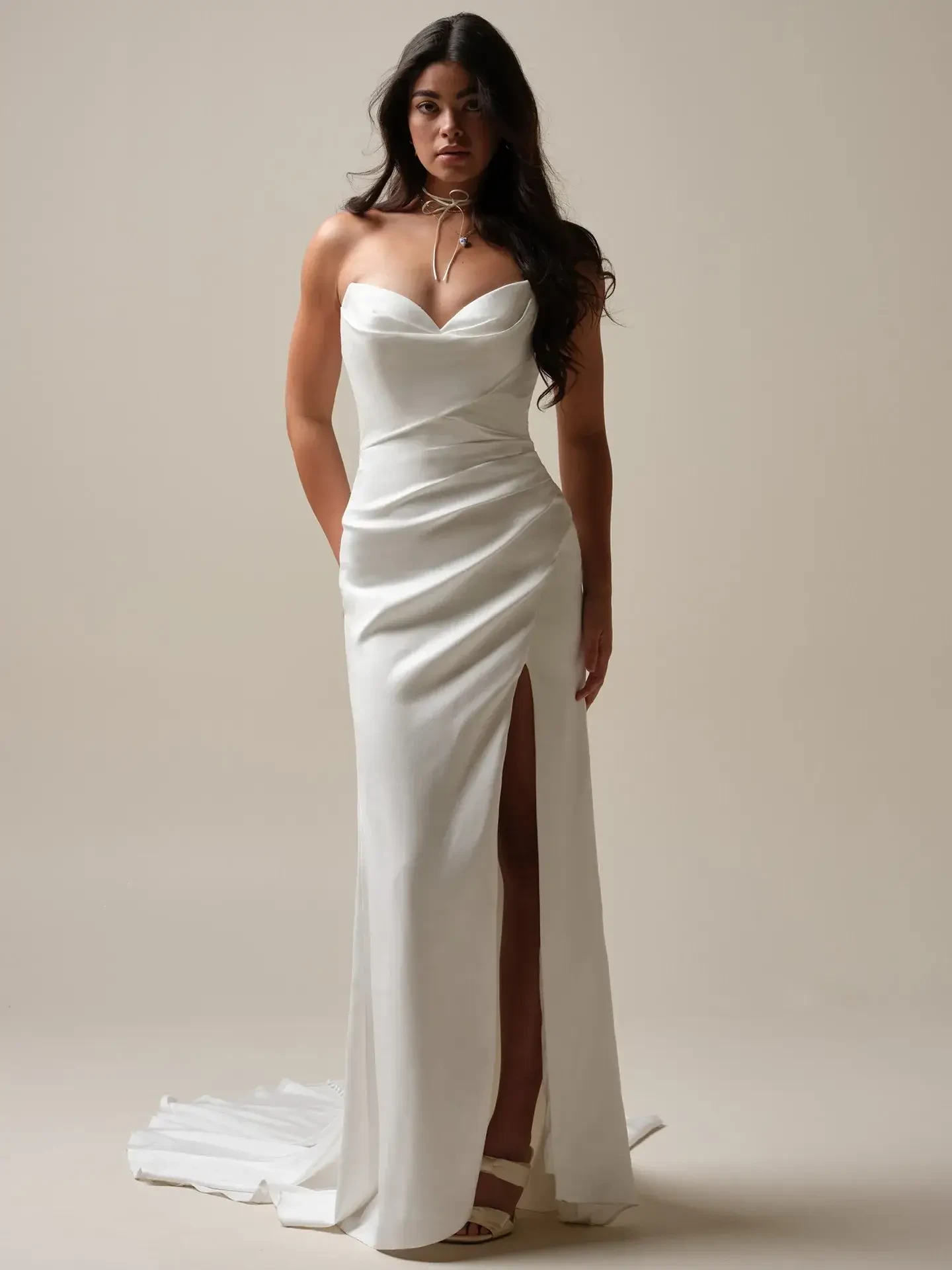 Elegant woman in a fitted white strapless gown with a thigh-high slit, wearing a delicate necklace. She stands poised against a soft beige backdrop.