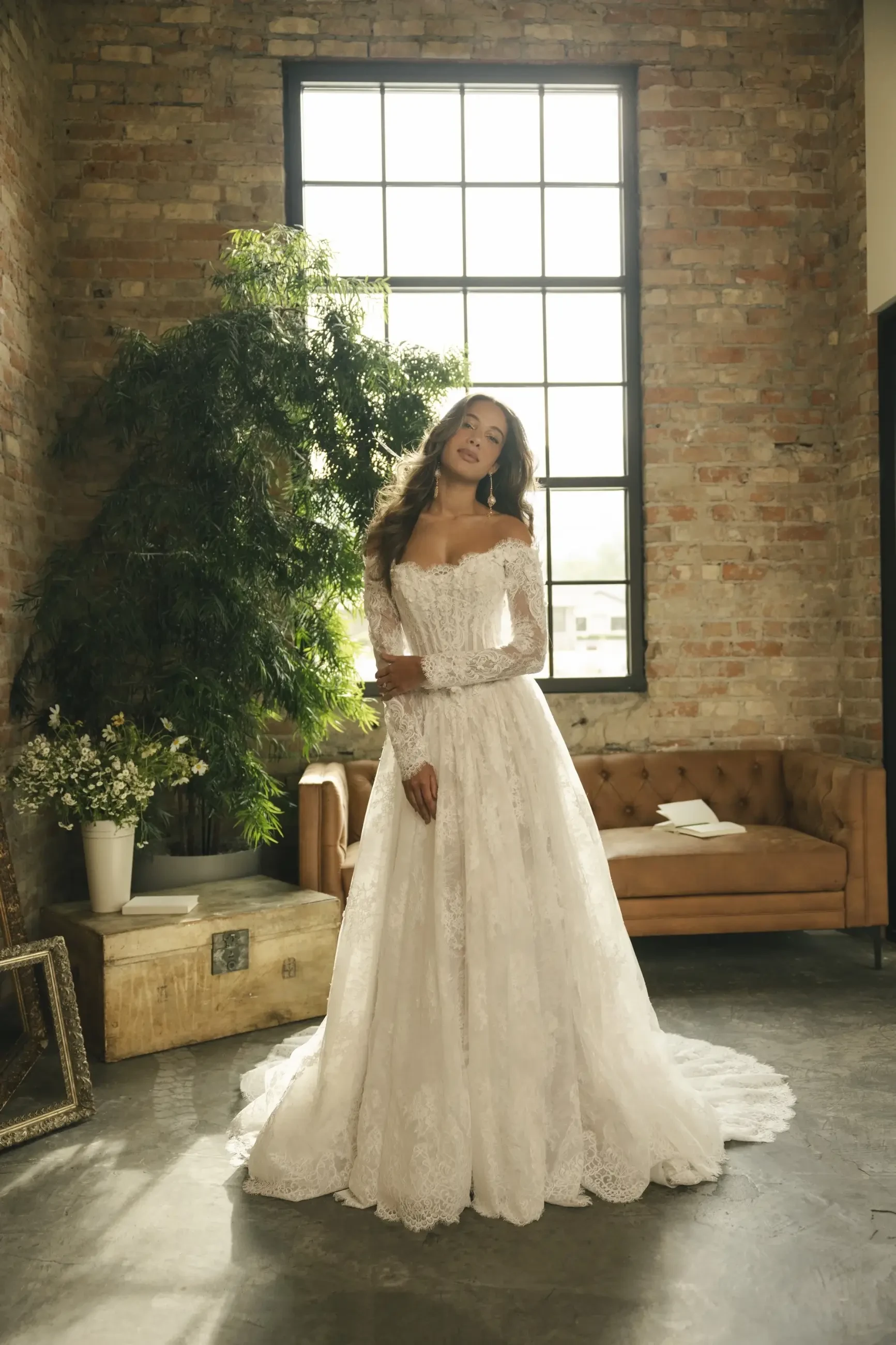 A woman in a lace wedding dress stands in a rustic room with brick walls, soft light from a large window, and a brown couch, evoking a romantic ambiance.