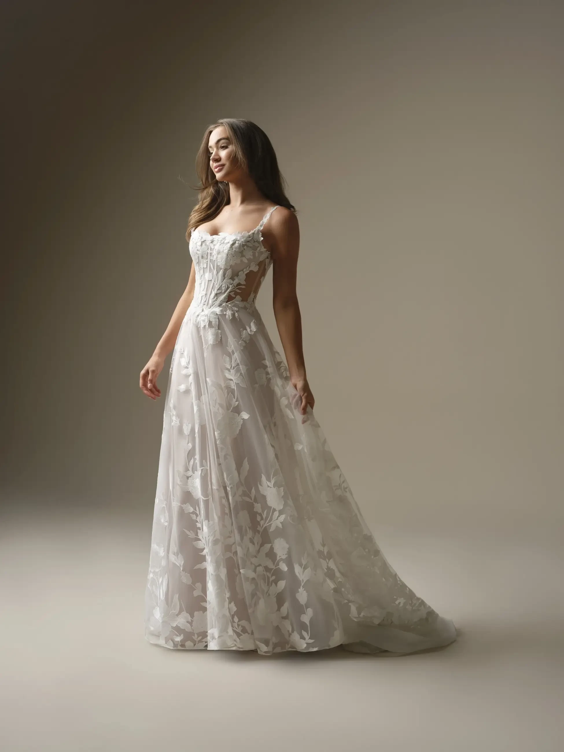 A woman wearing a beautifully detailed, flowing wedding gown stands elegantly against a neutral background. The dress features intricate floral patterns and delicate straps.