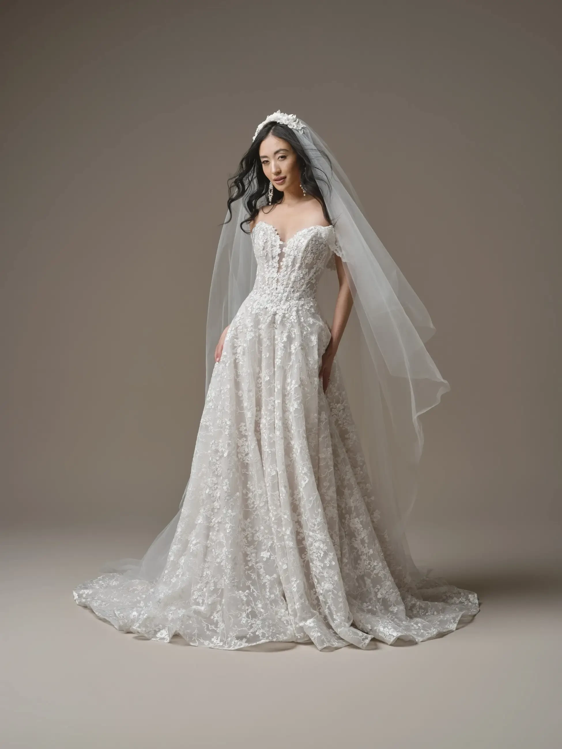 A bride in an elegant, lace off-the-shoulder wedding gown with floral patterns, complemented by a long veil. She stands gracefully against a neutral backdrop.