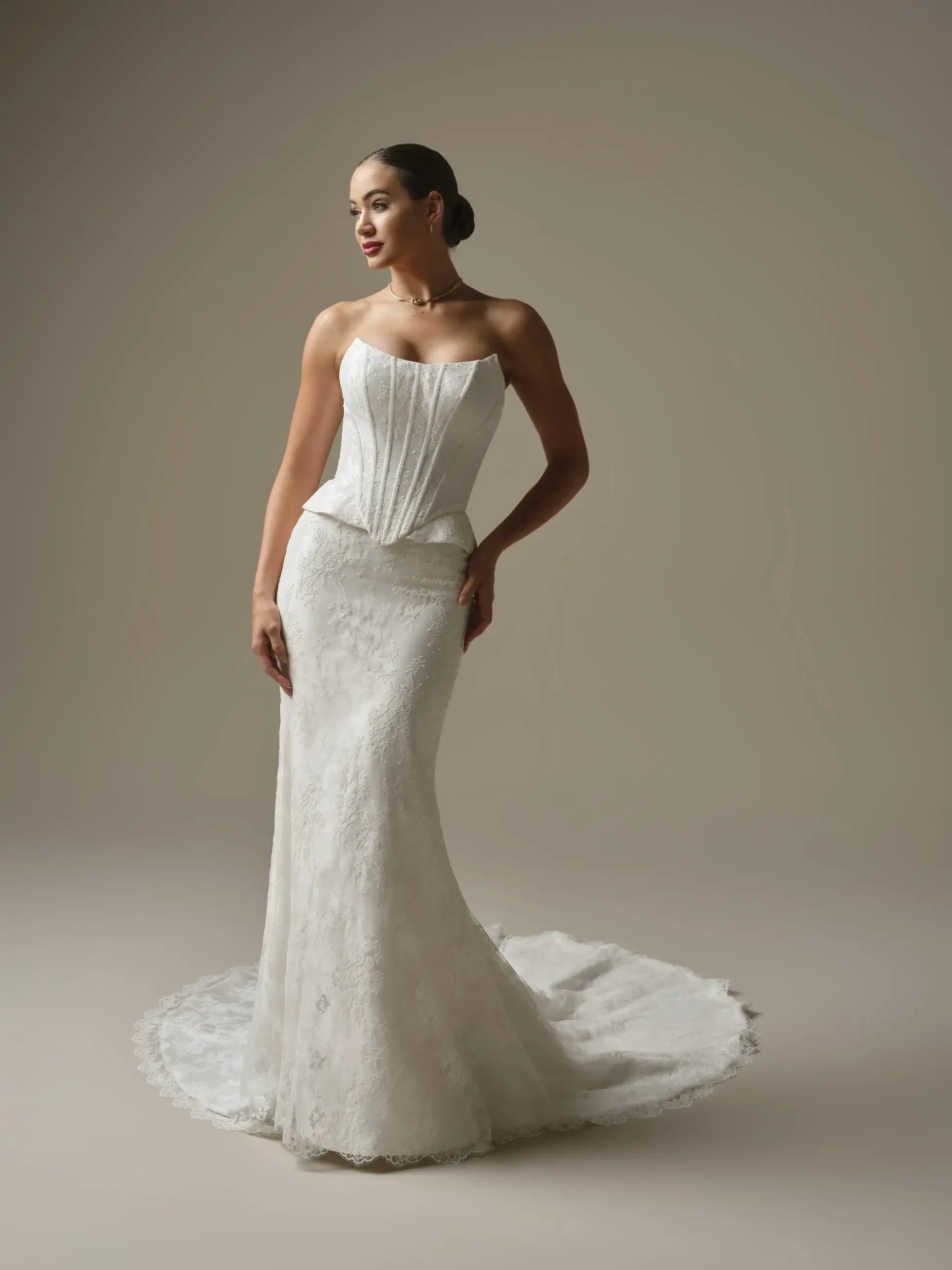 A woman in an elegant, form-fitting white wedding gown with a corset bodice and lace train stands gracefully against a neutral background. She exudes poise and confidence.