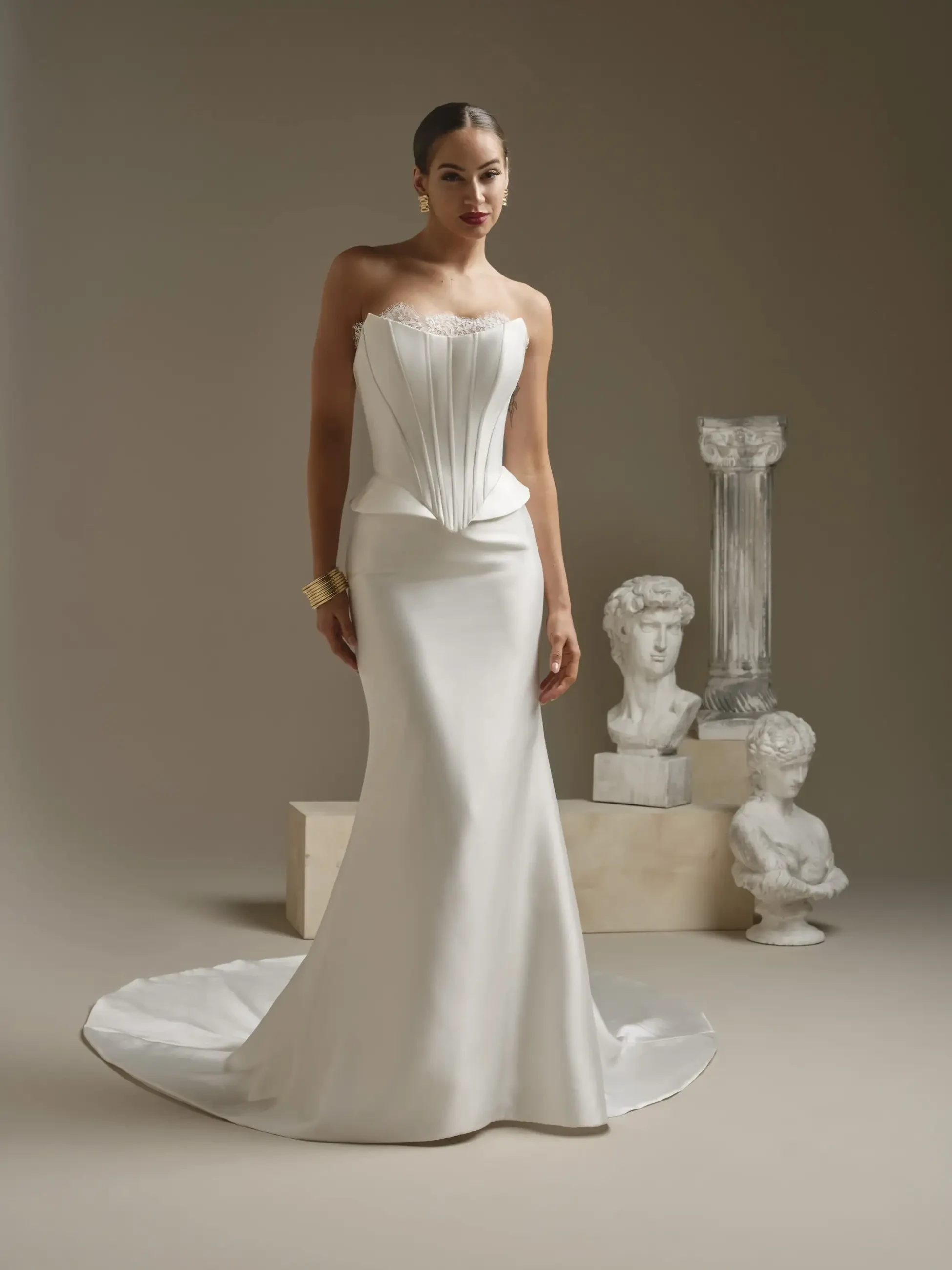 A woman in an elegant white strapless gown with a pleated bodice stands confidently. Behind her are classical-style busts and a tall column, exuding a timeless, artistic vibe.