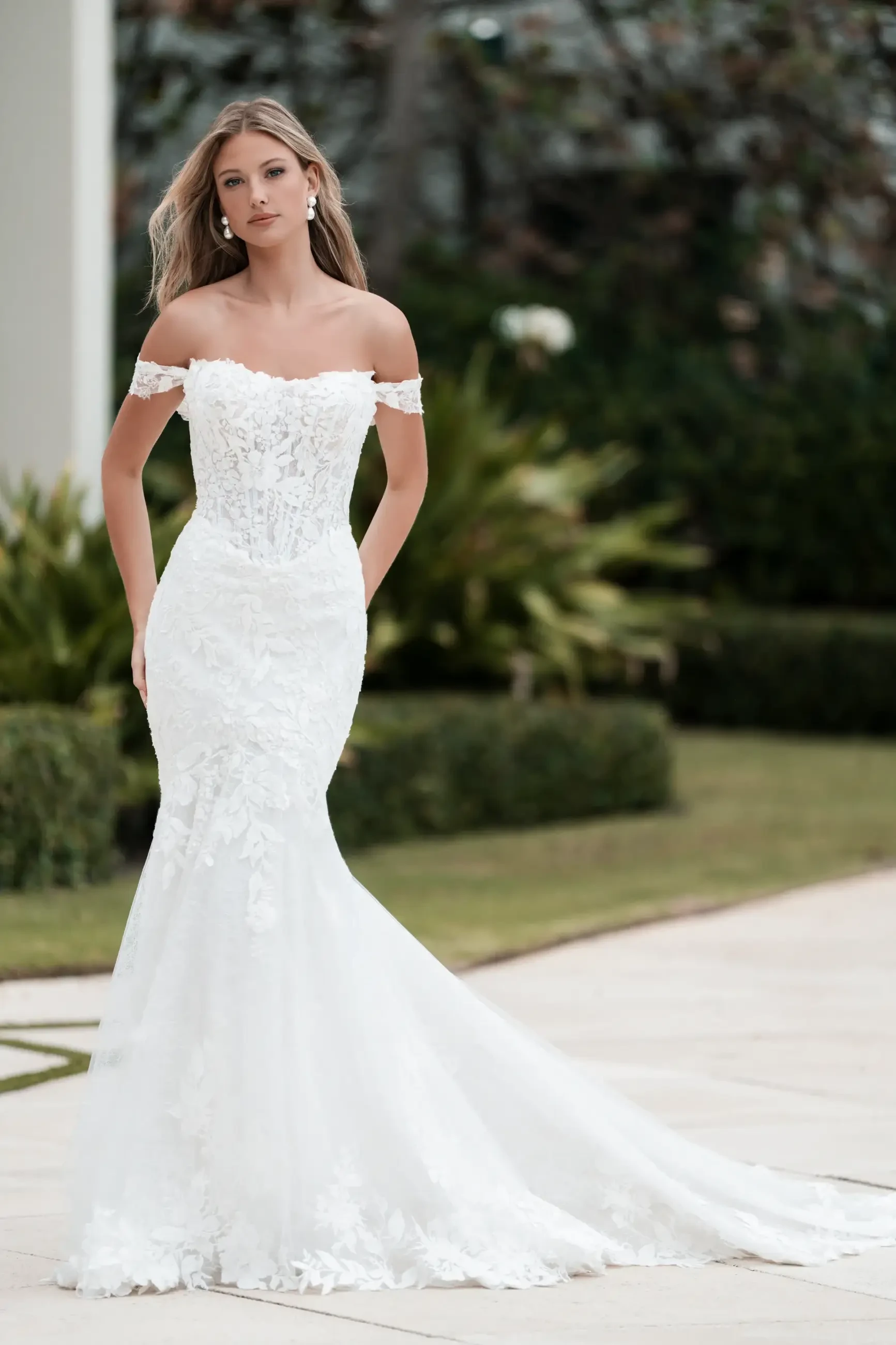 A woman in an elegant white off-the-shoulder wedding dress with floral details, posed outdoors in a garden setting.