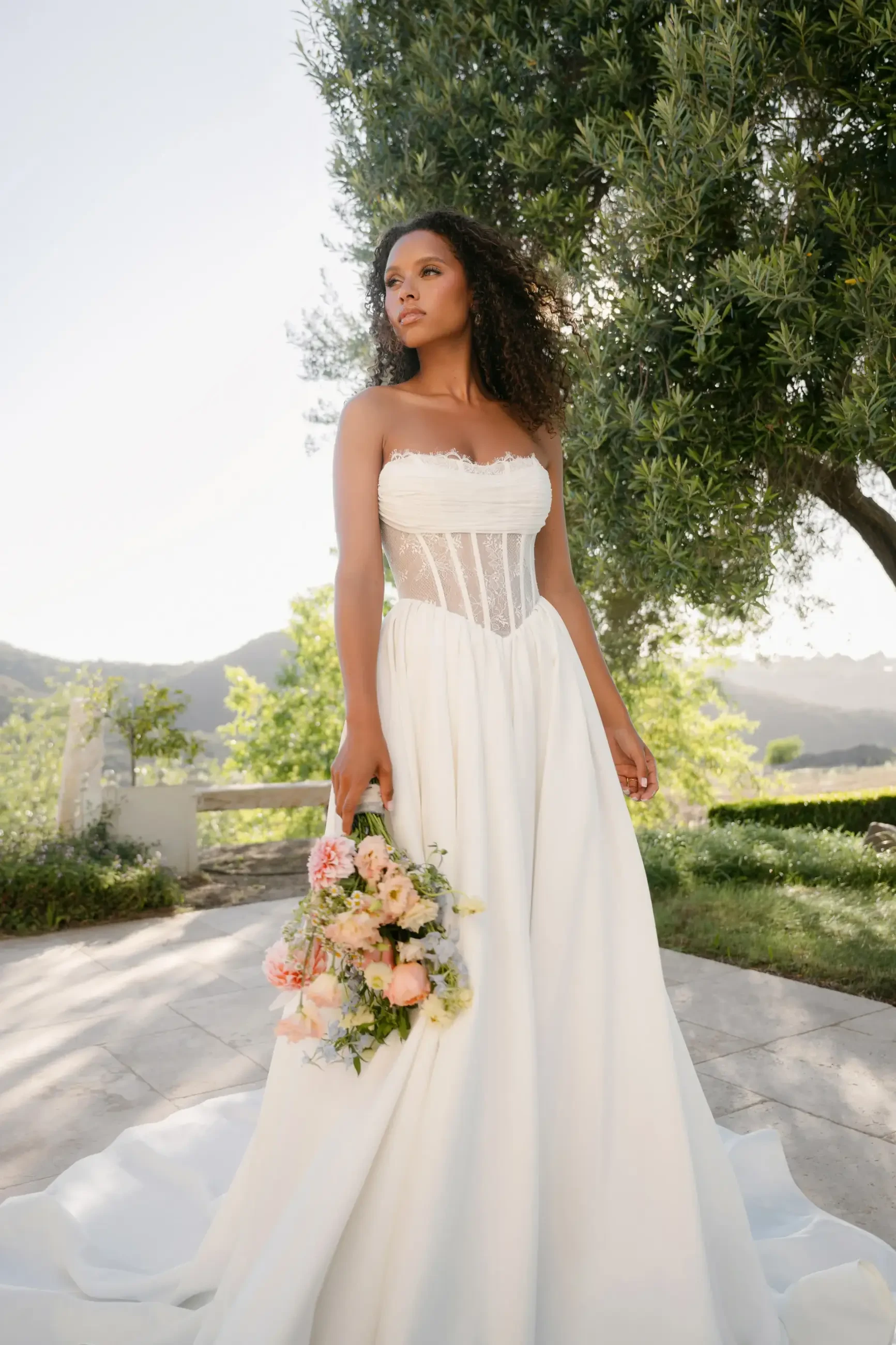 A woman in a strapless white gown stands outdoors, holding a bouquet of flowers. She's surrounded by greenery and mountains in the background.