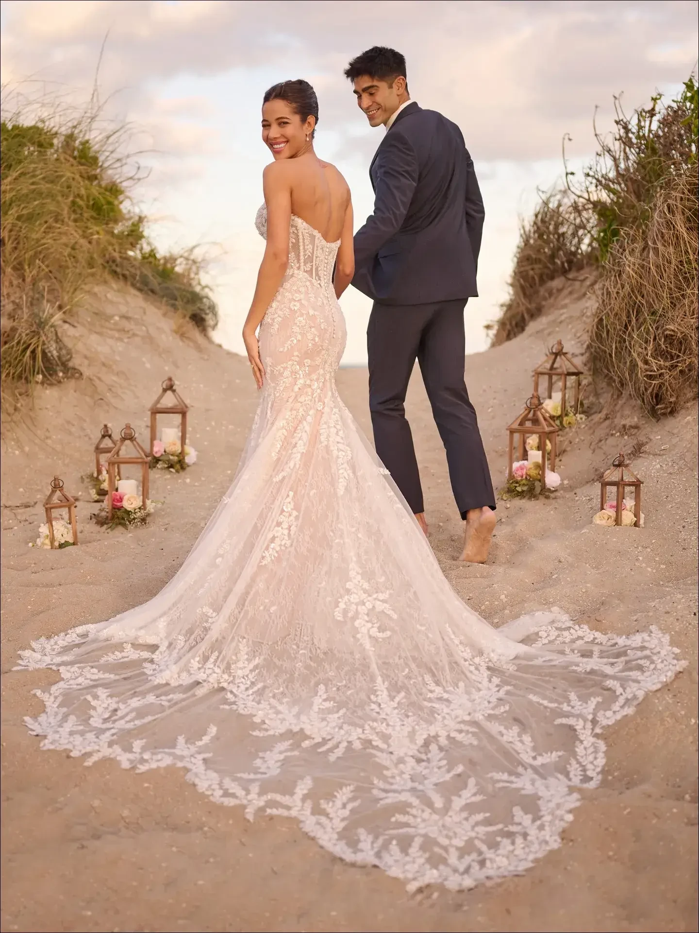 A couple stands on a sandy path adorned with lanterns; the woman is wearing a beautiful lace wedding dress, and the man is dressed in a dark suit.